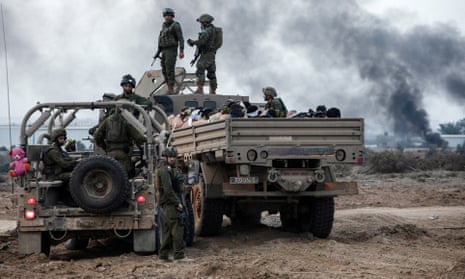 With black smoke billowing behind them, two soldiers in tactical gear stand on the cab of a pickup truck loaded with seated people, alongside a jeep full of other soldiers in tactical gear.