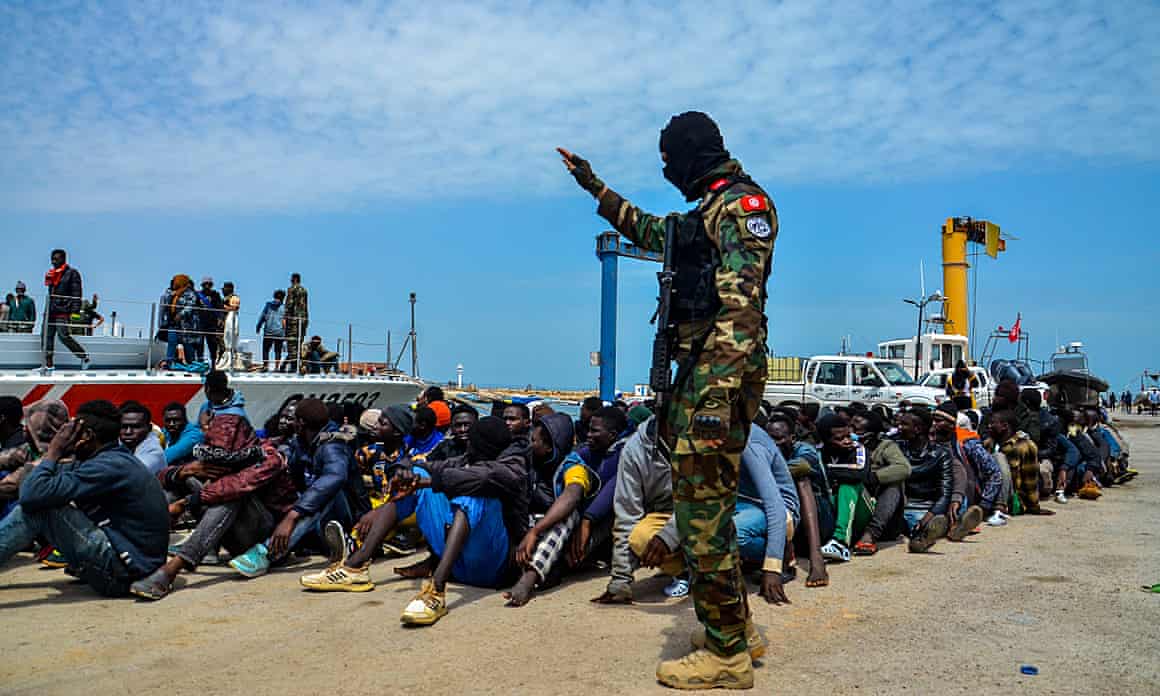 African migrants sit on the floor as a soldier stands nearby