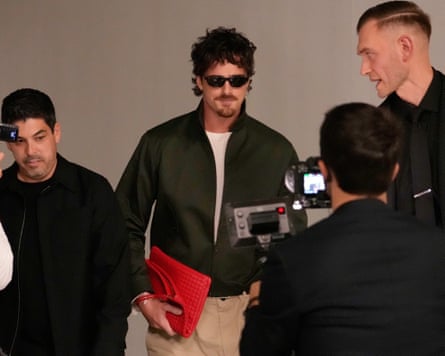 Elordi, wearing sunglasses, holds a red clutch while walking past photographers at a fashion show