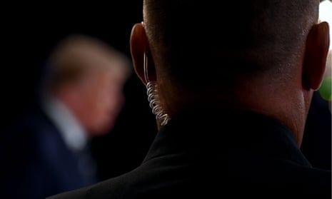 Close up image of the back of a man's head who is wearing a communication device in his left ear. Blurred, in the background, is the figure of Donald Trump.
