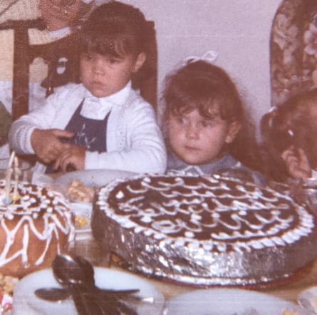 Two little girls stand at a table looking at a two big round shallow chocolate cakes, one with tinfoil wrapped around the sides and white piped patterns on the top