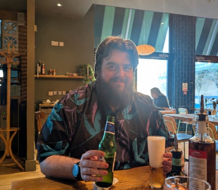 Chris sits at a wooden table with a bottle of beer. He has a big, bushy beard and wears a brightly patterned shirt.