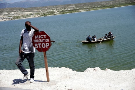 A man leans on a sign as a group of people sail in a rowboat at the border of Malpasse, Haiti.