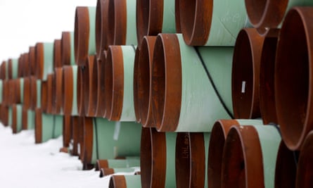 In snow and under a white sky, a long stack three high of wide metal pipes, painted light green with rusted ends.