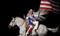 woman on a white horse holding an american flag and wearing a red, white and blue jumpsuit and a white cowboy hat
