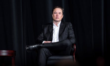 Elon Musk dressed in dark clothing, sitting looking at the camera against a dark background