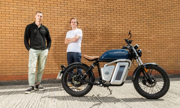 Two young men stand next to a retro-looking small motorbike with a leather saddle and black wire wheels, with a battery assembly where the motor would be