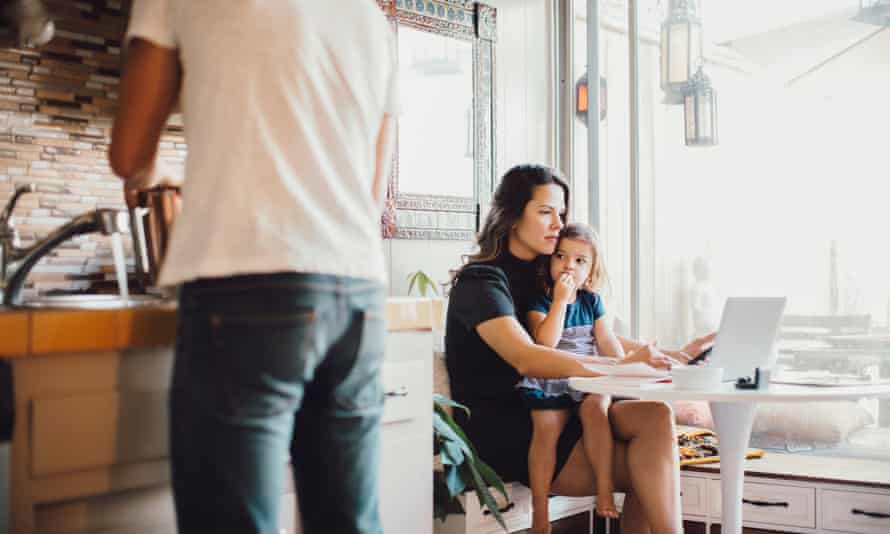 Woman holding toddler while working on laptop with male at sink in foreground.