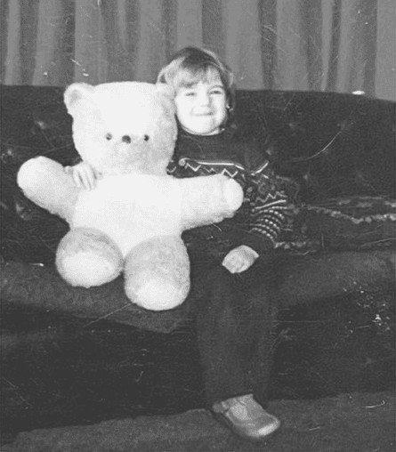 Jenni aged about 3, sitting on a sofa grinning with a giant teddy