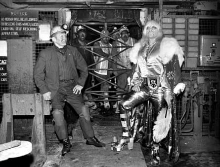 Flamboyant blonde wrestling star in 1970s style glam clothes and high heels pictured in a mine with his father and other men in traditional mining clothes
