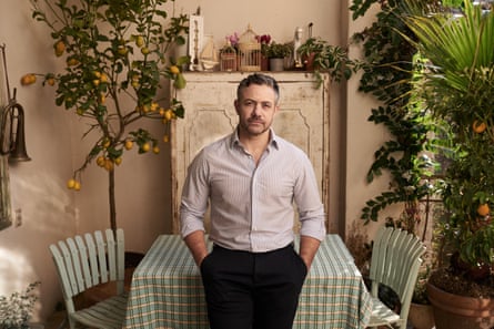 Wearing a light shirt and dark trousers, he leans against an outdoor dining table with his hands in his pockets, a lemon tree in the background