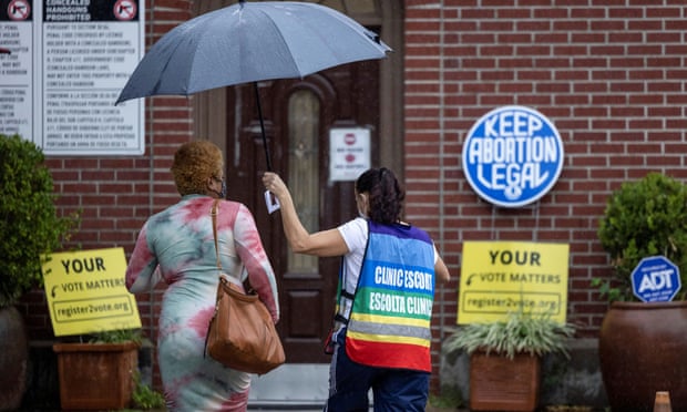 A woman in a rainbow colored vest that reads 'clinic escort' holds an umbrella over another woman's head as they both walk into a brick building. Just next to the door is a sign that reads 'Keep abortion legal'.