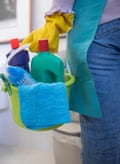 A woman wearing an apron and rubber gloves and jeans stands in a bathroom holding a blue bucket full of chemicals and facilities for cleaning