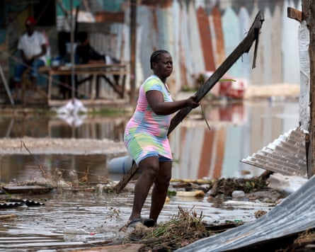 A woman standing in flood water carries a long plank next to a damaged building partly made of corrugated iron