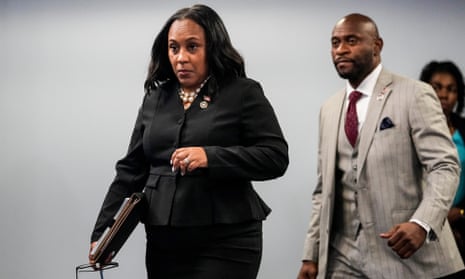 a woman in a black suit walks ahead of a ma in a grey striped suit with a red tie