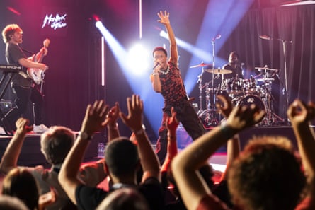 A man on stage holding a microphone and waving his arm above his head as people in the audience wave back.
