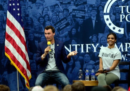 A man and woman sit on chairs on stage in casual clothes next to a US flag, the man holding a mic and talking