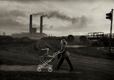 A woman pushes a pram across a muddy site, smoking factory chimneys behind, in Consett