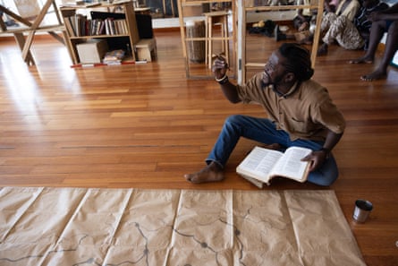 An African man sits on a wooden floor by a large bit of paper, gesturing as he holds a book