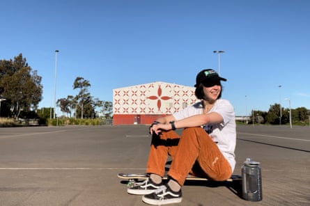 Seorang wanita dengan topi hitam, T-shirt putih dan celana oranye duduk di longboard.
