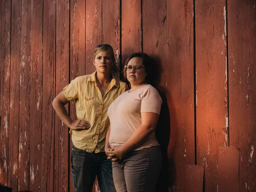 Two women, one in a short-sleeved yellow button-down shirt and black jeans, the other in a pink T-shirt and gray jeans, lean against the outside wall of a red barn.
