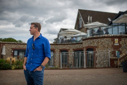 James Nye stands in front of the pub building and its outdoor terrace; he wears a bright blue linen shirt with jeans and has short light brown hair. He stands with his hands in his pockets