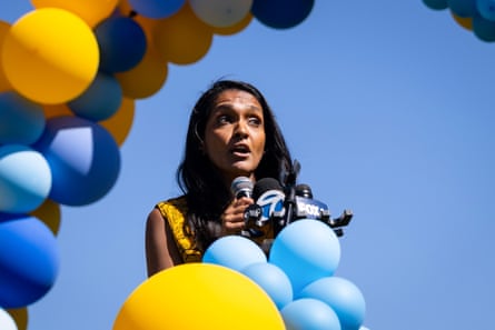 A woman speaks into a microphone with bunches of yellow and blue balloons around her.