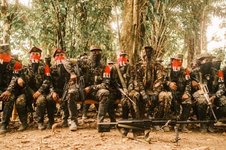 A group of fighters in combat gear and with red and black bandanas covering their noses and mouths sitting in a jungle clearing