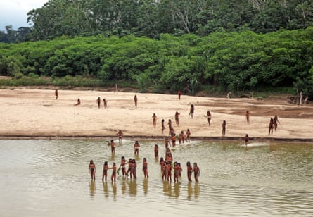 A group of Indigenous people stand in and on the banks of a river next to rainforest