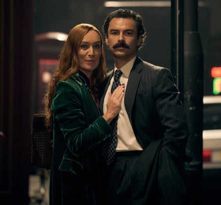 A man in a suit and tie stands with his arm around a woman in a green coat