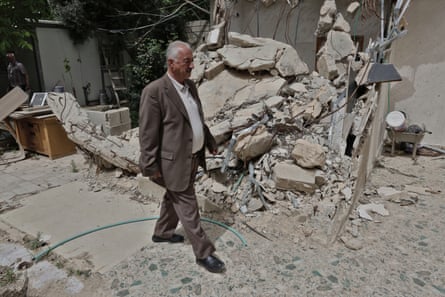 Fakhri Abu Diab walks by the rubble of his house. He has white hair and wears a dark grey suit with open jacket over a white shirt.