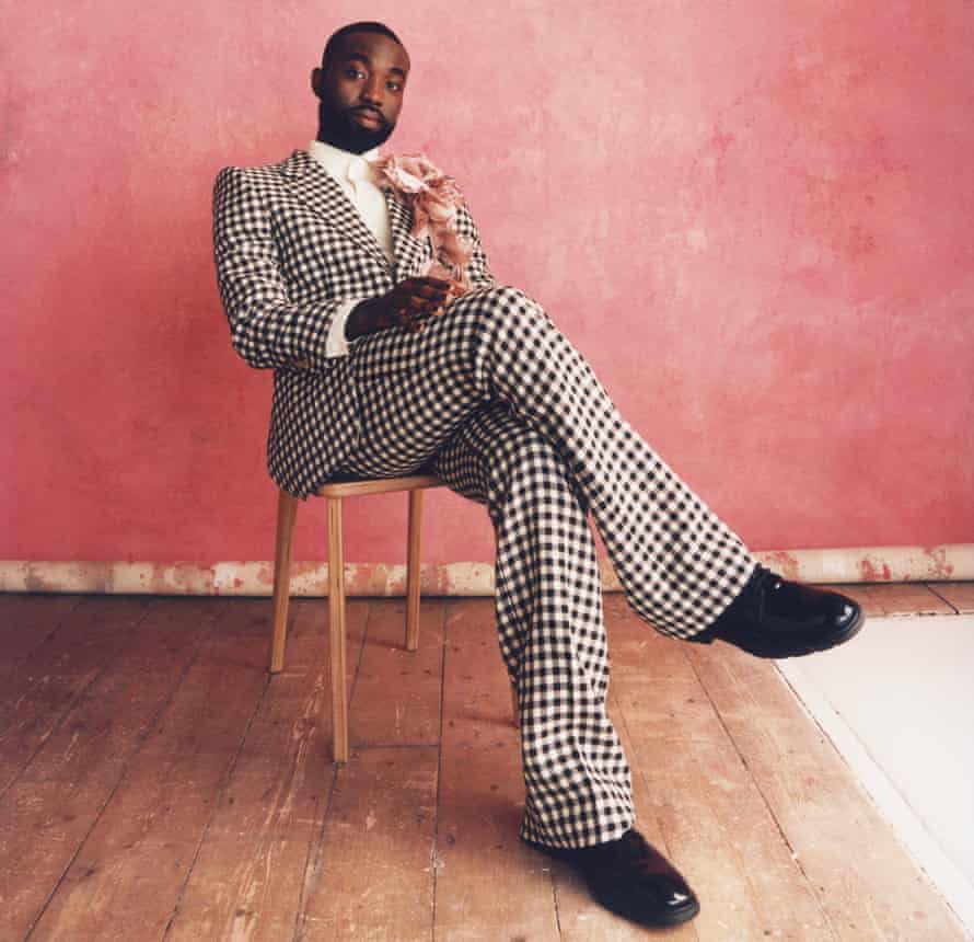 Actor Paapa Essiedu in black and white check suit, sitting on a dining chair, against pink background