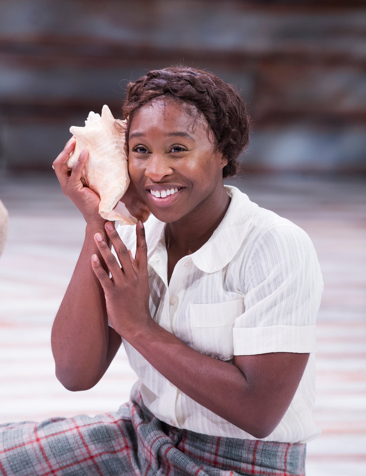 Cynthia Erivo sitting on the floor on stage smiling and holding a large seashell to her ear