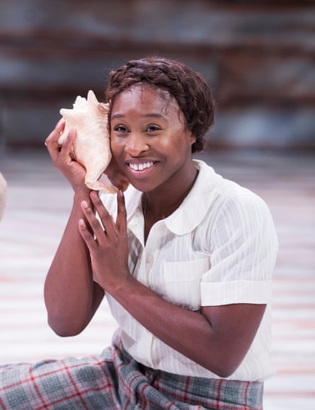 Cynthia Erivo sitting on the floor on stage smiling and holding a large seashell to her ear