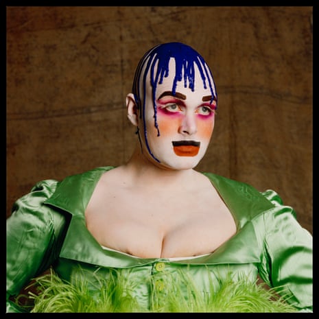 Leigh Bowery is photographed in a low-cut satin dress wearing heavy makeup and what looks to be purple paing dribbling down off his head