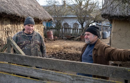 Gheorghe Boftea (left) and Mititelu Petrea standing in a muddy enclosure behind a wooden fence, with straw-roofed farm buildings on either side and a white house behind