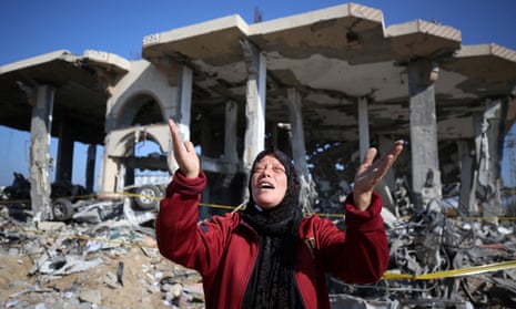 A Palestinian woman holds her hands up to the sky in front of a destroyed building and rubble