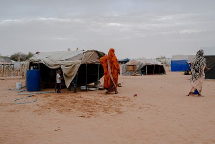 Two women wearing shawls draped over their heads stand on the sand next to tents.