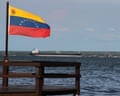 Red, yellow, and blue flag in wooden pier, with choppy water and very long ship beyond.