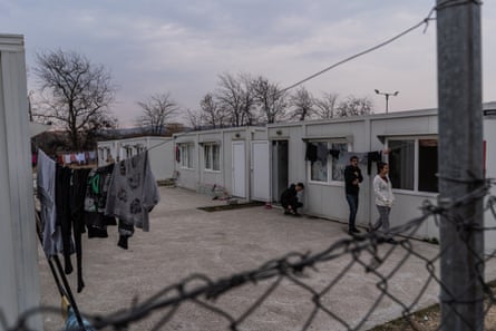 People can be seen in a courtyard next to a row of small buildings. There is washing on a line. A fence can be seen in the foreground