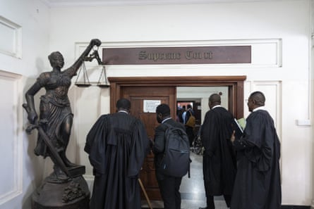 A group of men, some in legal gowns, pause in front of a door next to a statue of Justice.