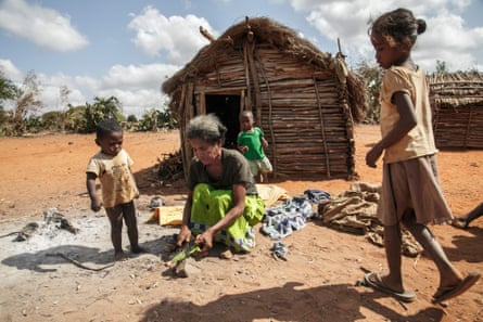A woman prepares food outside a wooden hut while three young children watch her.