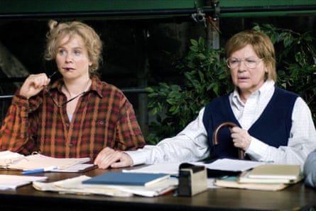 The two women sit at a table covered in files and papers, looking businesslike
