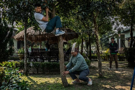 A man wearing a makeshift mask crouches next to a small tree. Another man has climbed halfway up the tree