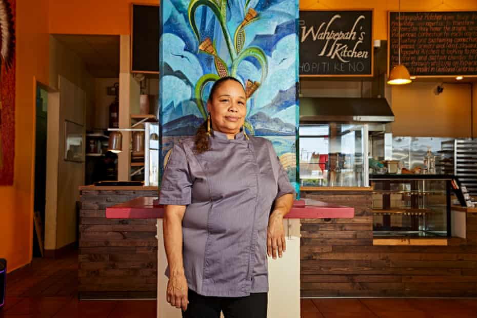 A woman in a gray chef’s coat stands in her restaurant, one arm propped on a counter. A sign behind her reads ‘Wahpepah’s Kitchen’.