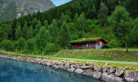 A Norwegian cabin by the water's edge; in the background is a heavily forested area of hills and mountains.
