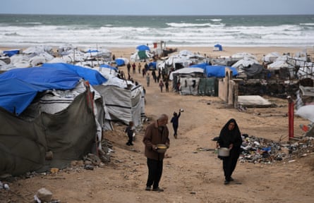 Two elderly people walk between tents pitched on a beach with the sea behind