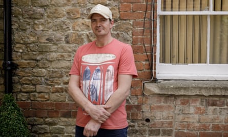 Charlie Rowley stands in front of a brick wall and window; he wears a light red T-shirt with surfboard and Volkswagen logos and a cream baseball cap
