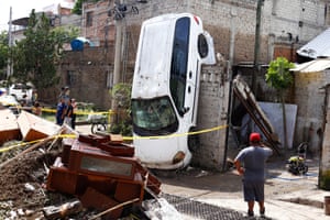 A man in a red baseball cap looks at a white car upended on to its nose against a wall amid other debris left by floods
