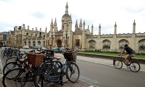 A cyclist in Cambridge rides past a bunch of parked bikes in the foreground and part of the university in the background.
